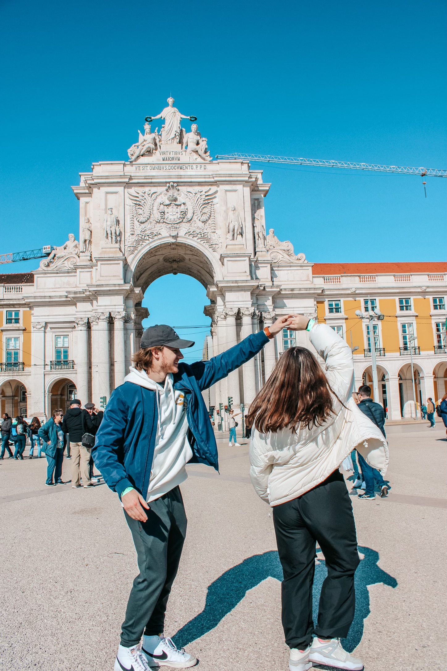 Woman in white jacket twirling with man in black baseball cap and blue jacket in front of Arco da Rua Agusta