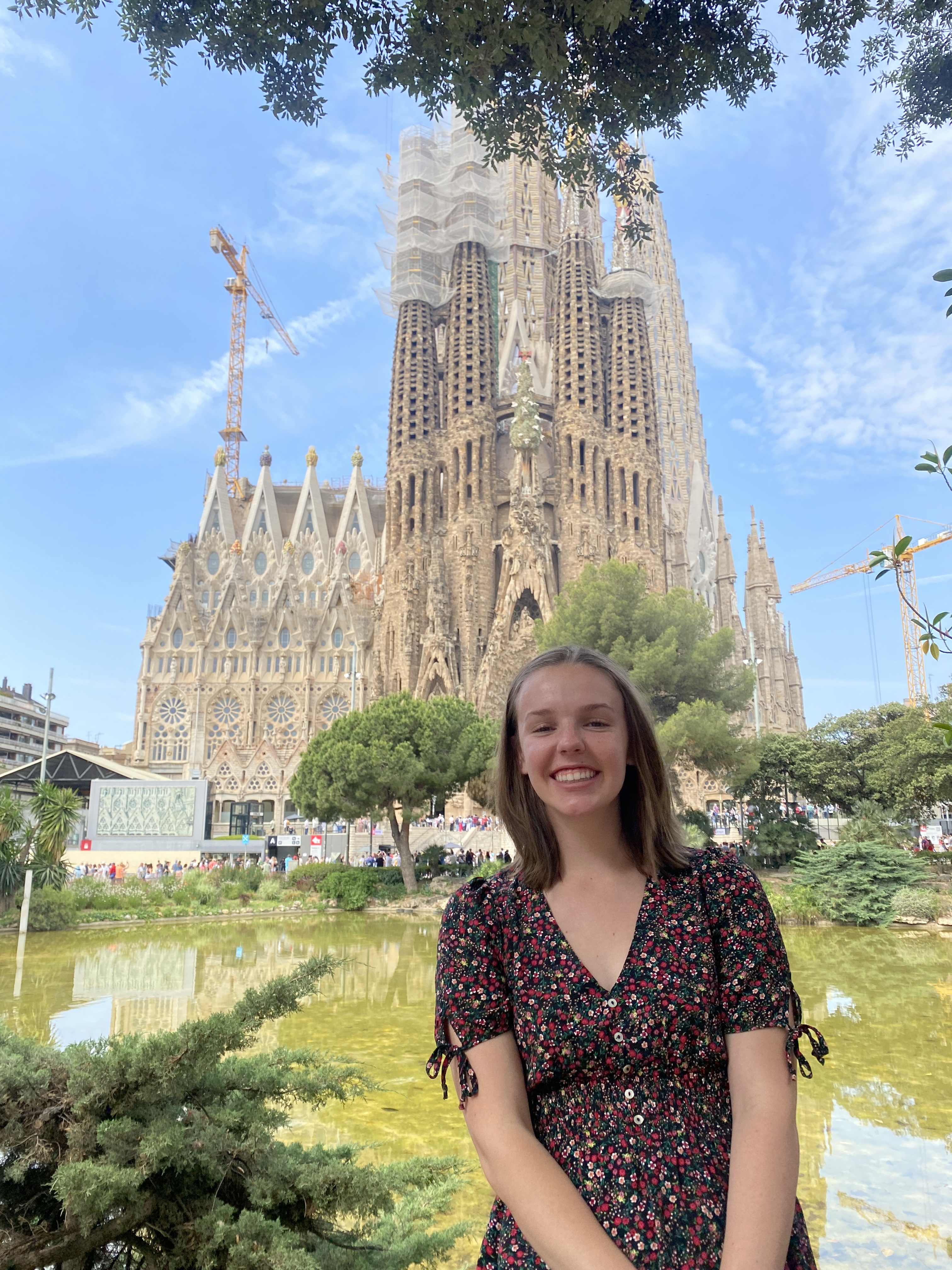 A person standing and smiling in front of La Sagrada Familia during the day at a park with trees and a pond in the background.