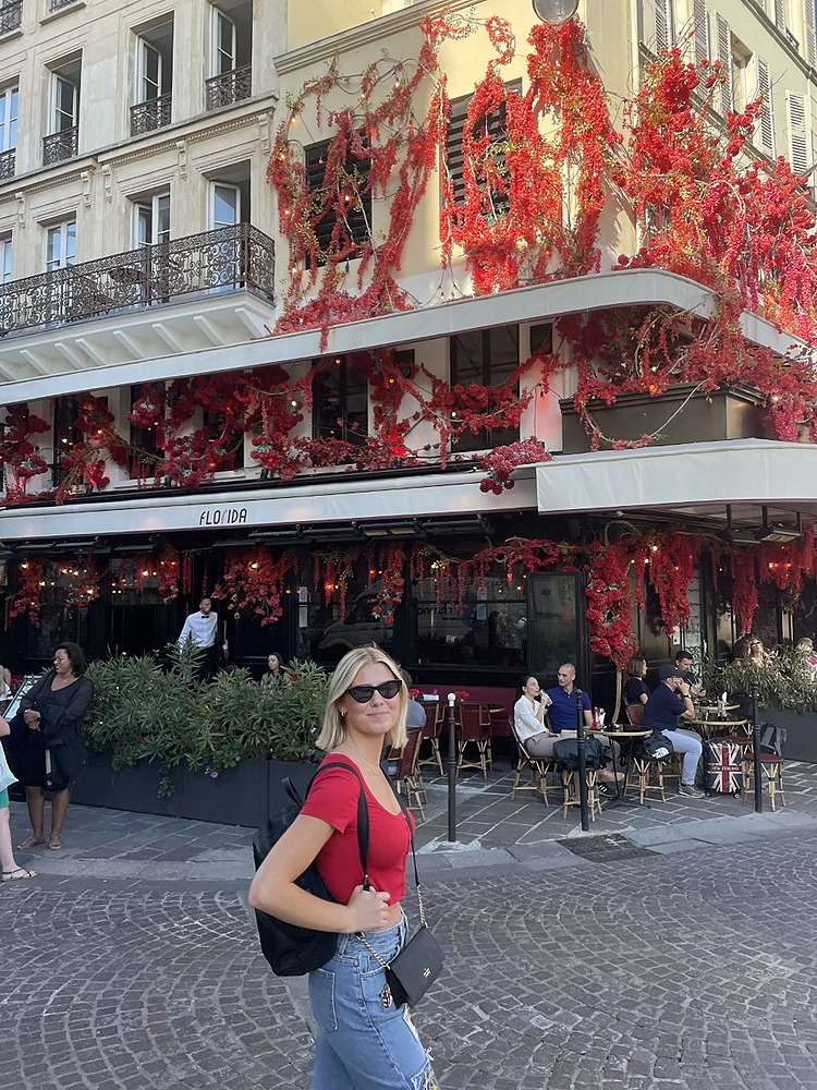 Blonde student wearing red tshirt and jeans posts in front of Paris store with matching red flowers.