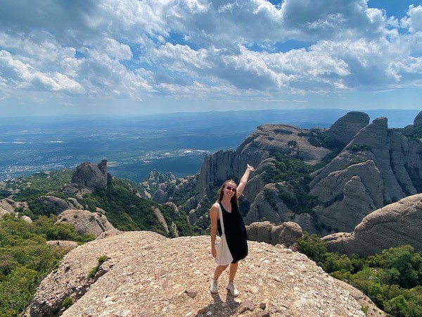 A person standing on a mountain while smiling and pointing their left arm at a mountain in the background.