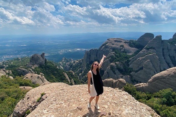 A person standing on a mountain while smiling and pointing their left arm at a mountain in the background.